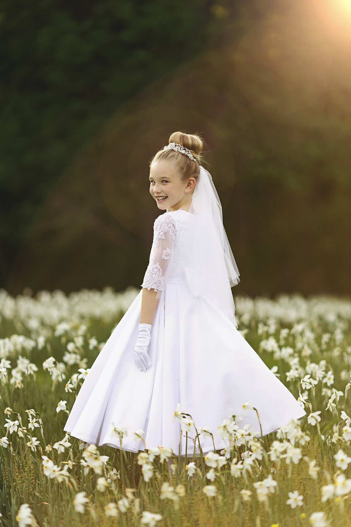 A young girl smiles over her shoulder while walking through a field of white flowers at sunset in a white dress