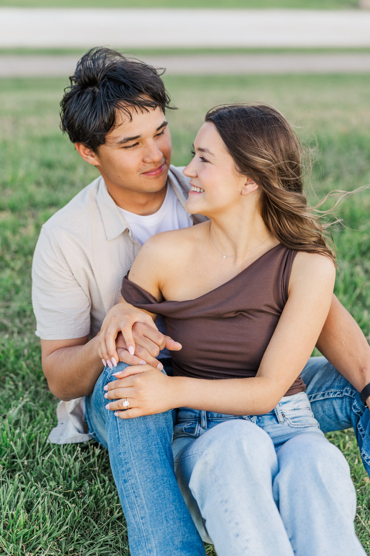 A joyful and candid moment between an engaged couple sitting in the grass during their Downtown Fort Worth engagement shoot. The bride-to-be’s diamond ring sparkles as they hold hands and share a loving smile, capturing genuine connection and romantic charm.