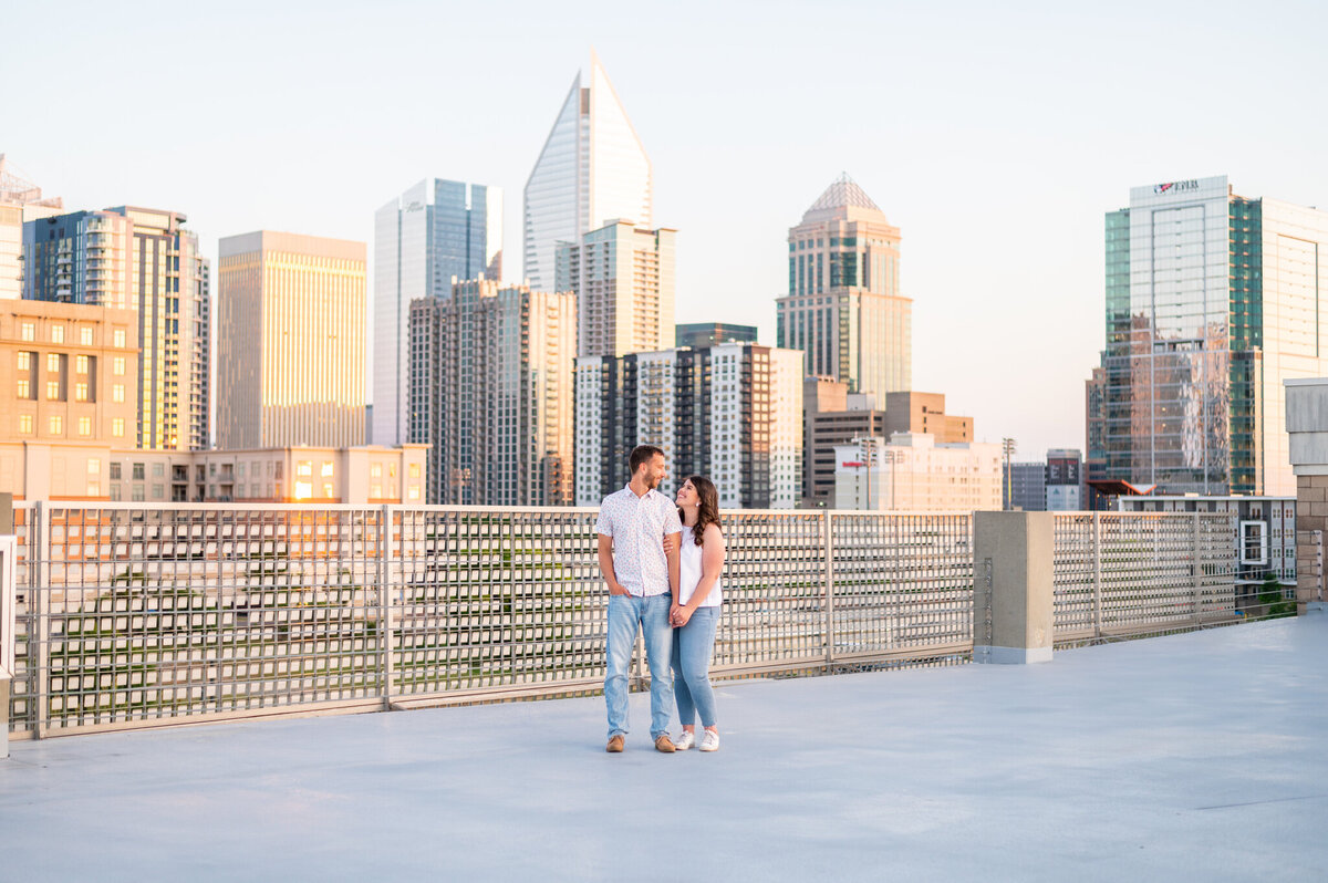 A woman holding the arm of her man with one hand and his hand with the other. Both are looking into each others eyes and smiling. Behind them is the Raleigh skyline.