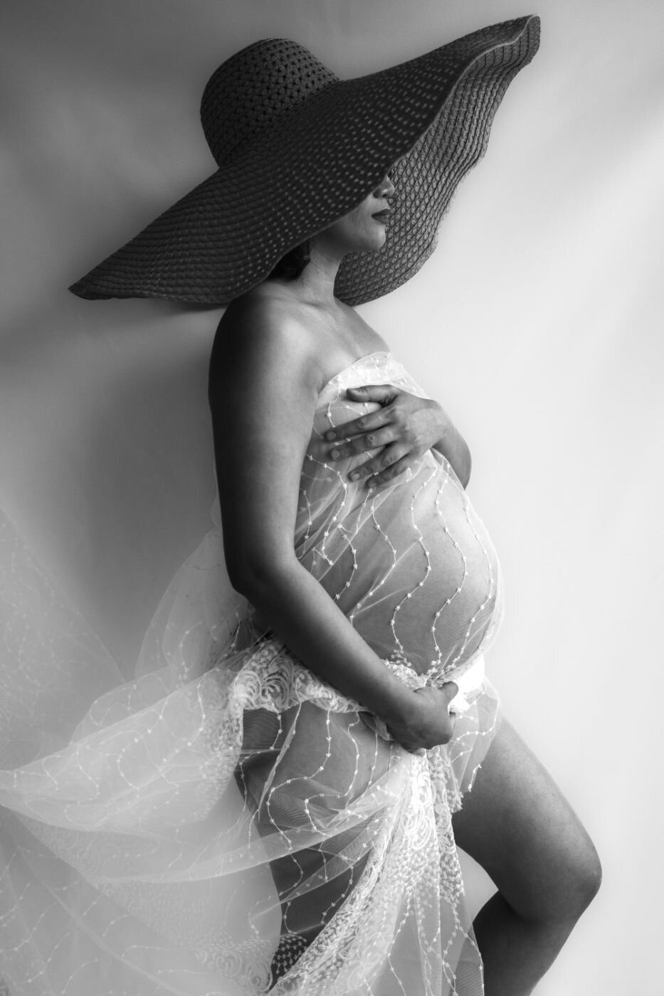 Side profile of expecting mom wearing a wide-brim hat holding her lace wrap across her chest and tenderly embracing her belly.