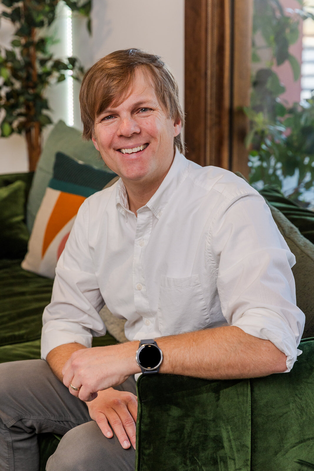 Man wearing white button up shirt sitting on a dark green sofa one arm on the handler smiling at the camera during portraits in Buckhead studio with Laure Photography