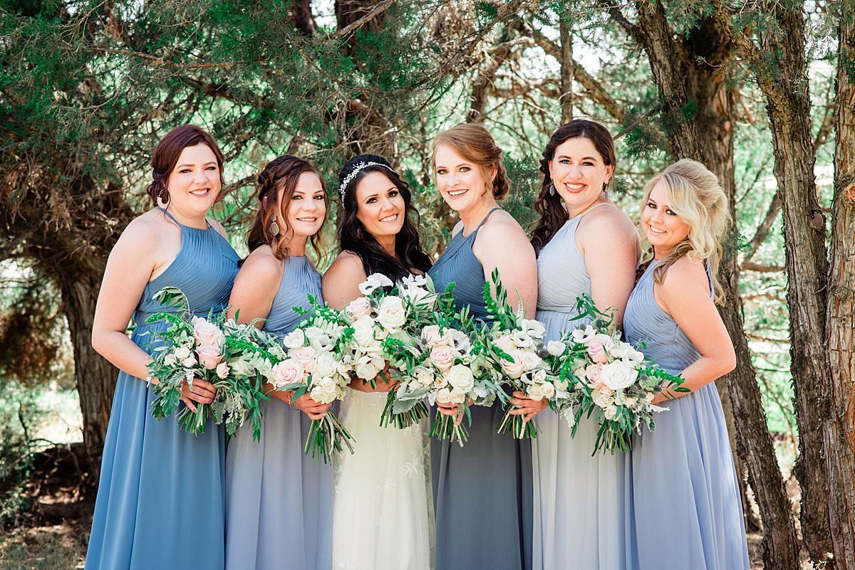 Bridesmaids in soft shades of blue dresses carrying neutral colored bouquets and smiling at the camera alongside the bride