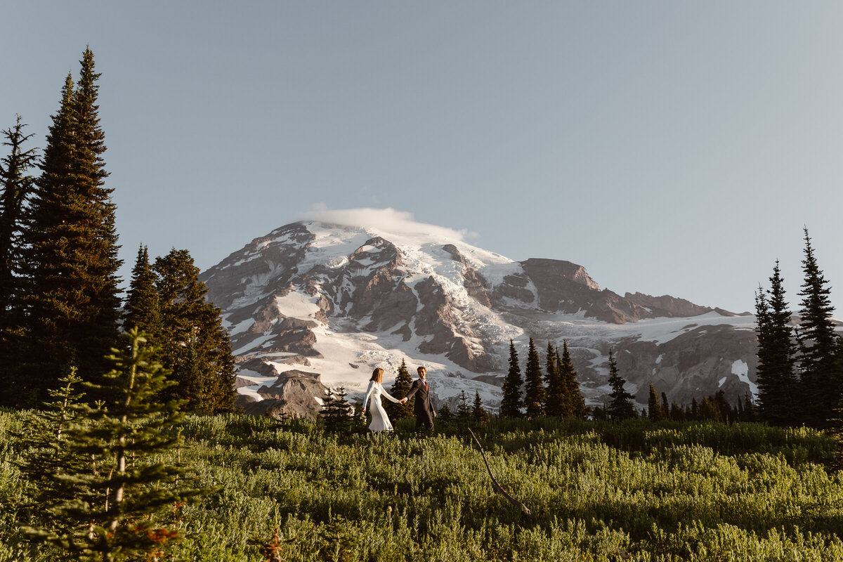 Man in suit leads woman in wedding dress along trail in front of Mount Rainier at sunrise