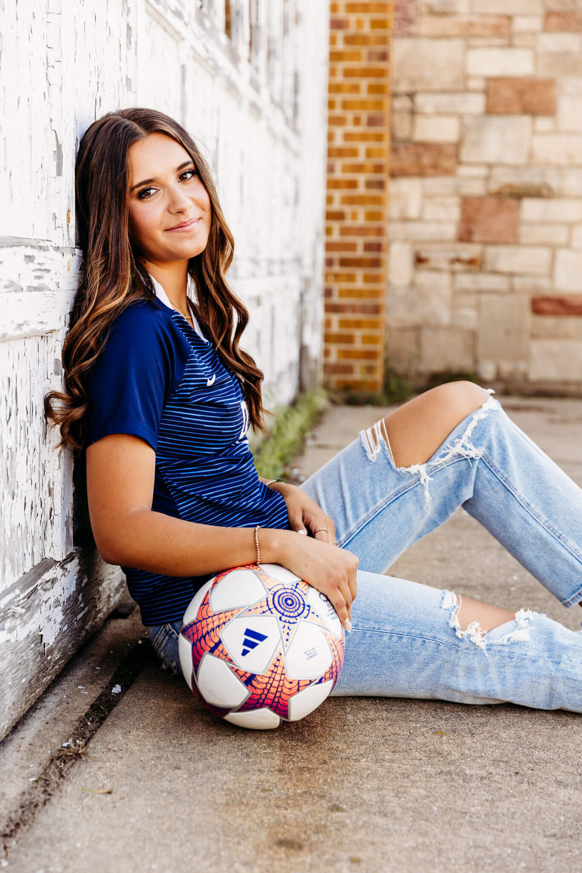stunning girl from Oshkosh West High School sitting against a rustic garage door with her arm on a soccer ball