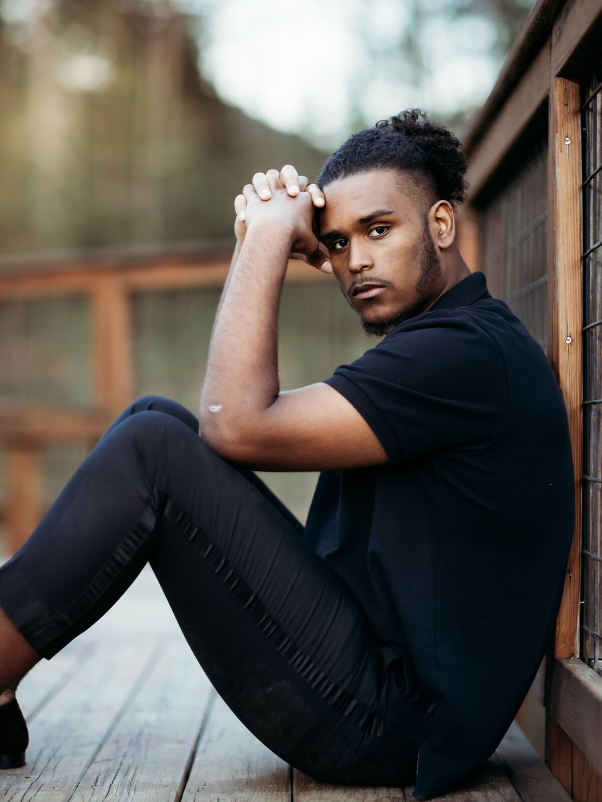 Boy in black shirt and pants sitting on a deck leaning against a post looking thoughtfully at the camera.