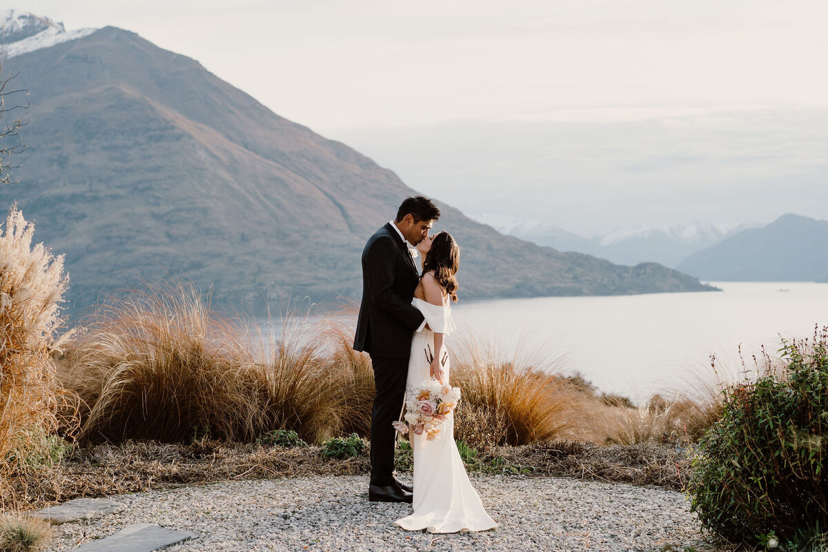 The Lovers Elopement Co - bride and groom kiss on lake side with Cecil Peak in background - Queenstown New Zealand