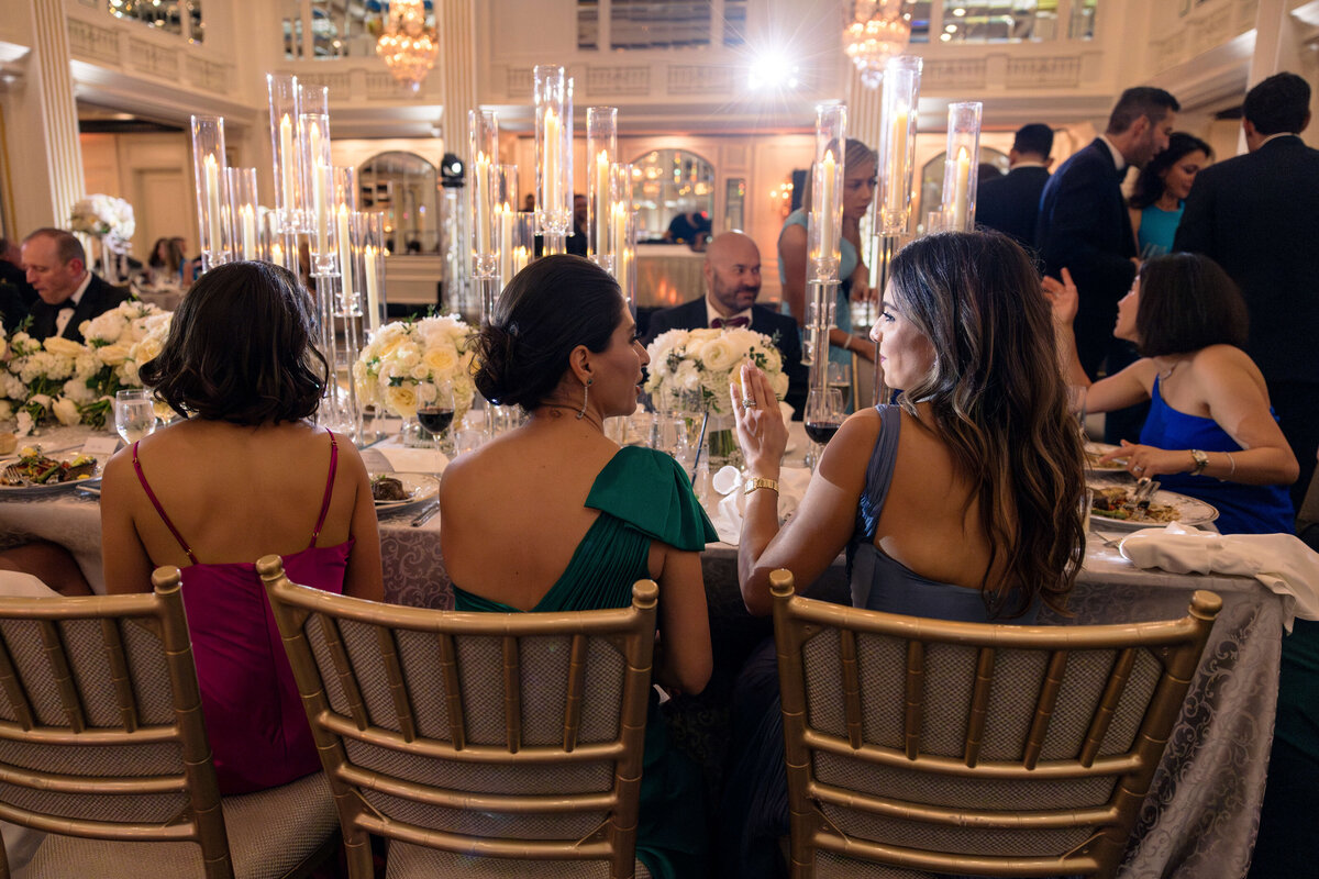 Guests in elegant attire sit at a banquet table adorned with tall candles and white floral arrangements, engaging in conversation. The room is lavishly decorated with chandeliers and ornate details, creating a sophisticated ambiance.