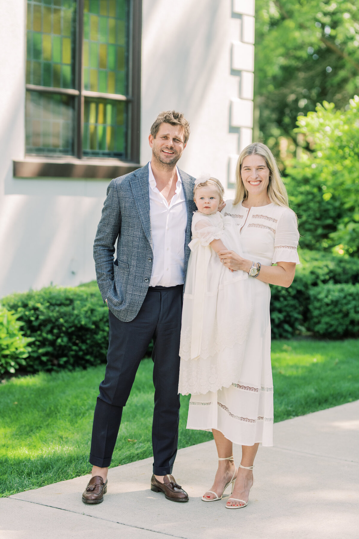 Elegant family of three in timeless attire, standing outside a historic Oakville church following their daughter’s baptism