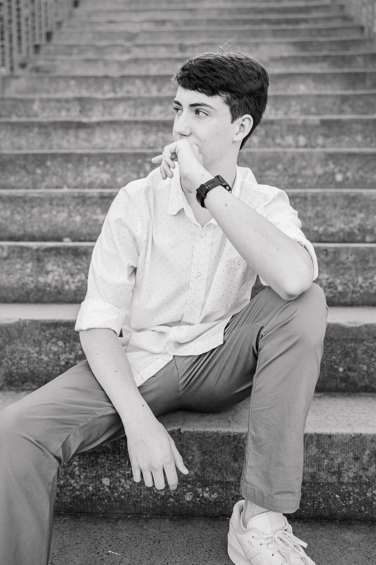 A young man sits on outdoor concrete steps, captured expertly by a senior photographer in Fayetteville, NC. Wearing a light-colored button-up shirt, dark pants, and white sneakers, he rests an elbow on his knee and his chin on his hand, gazing to the side in this striking black-and-white image.