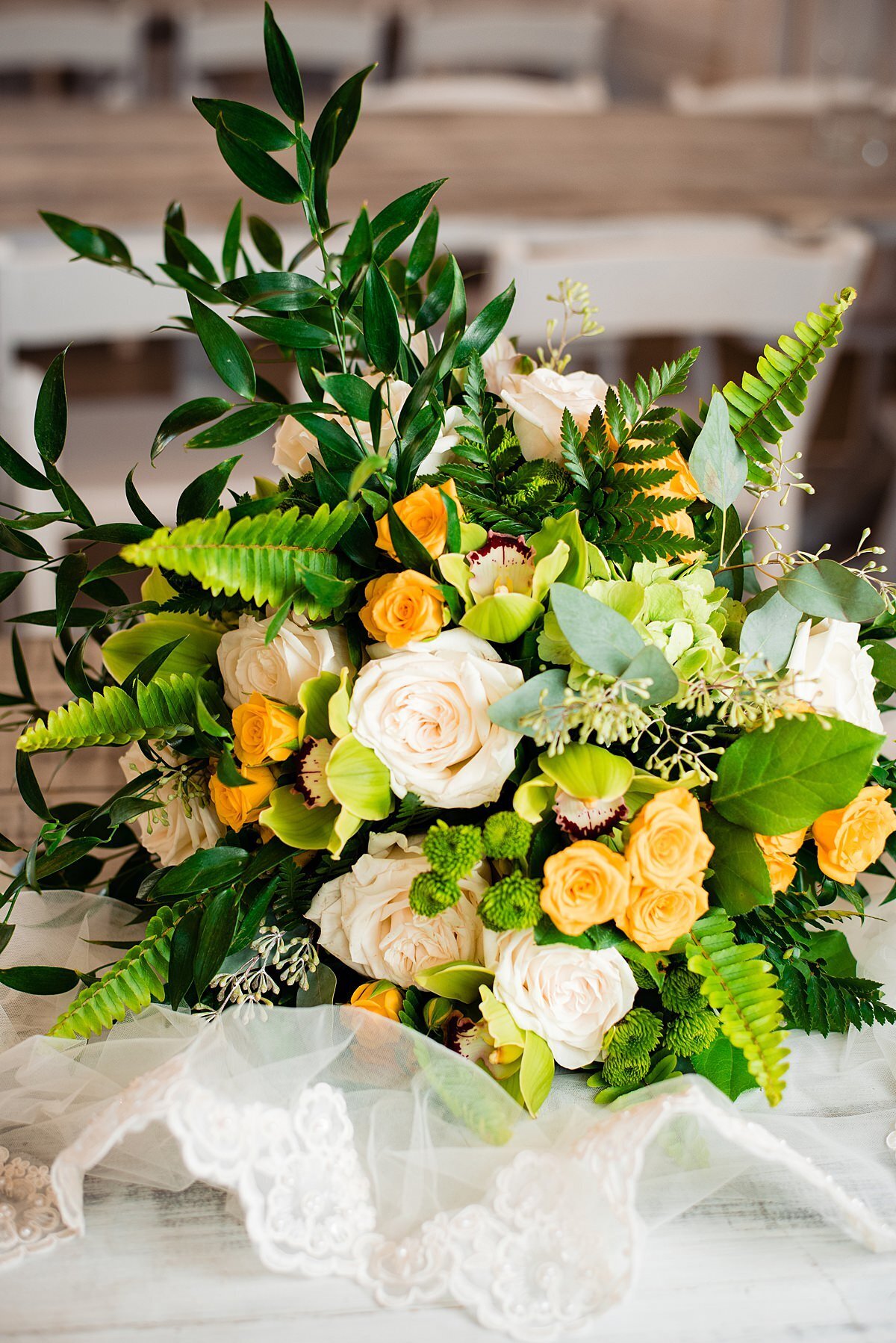 A large cascade bridal bouquet  with white roses, yellow tea roses, fern and greenery sits on a table with the bridal veil edges in lace.