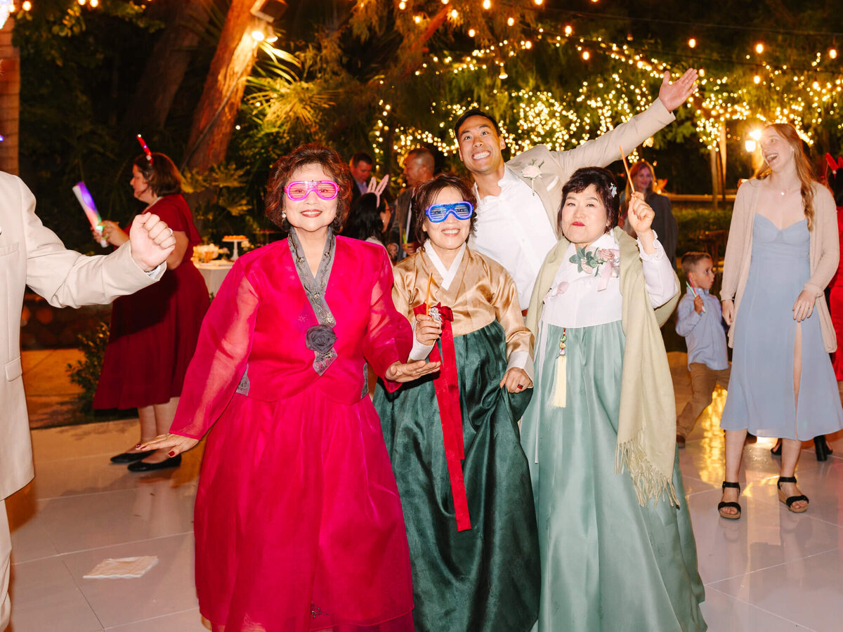 A joyful group at a festive event, wearing traditional Korean hanboks and colorful glasses, dance under twinkling lights.