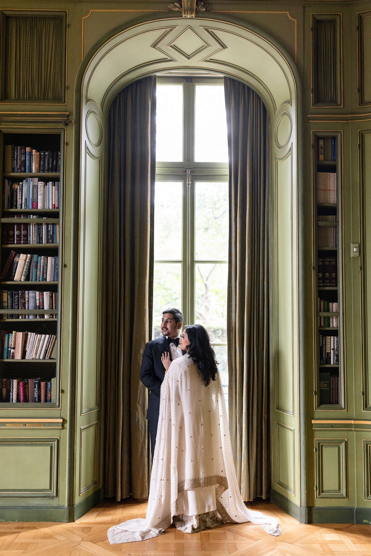 A couple stands by a large window in an elegant room with green walls and bookshelves. The woman, draped in a white cape, faces the man, who wears a black suit. They stand close together, gazing at one another.