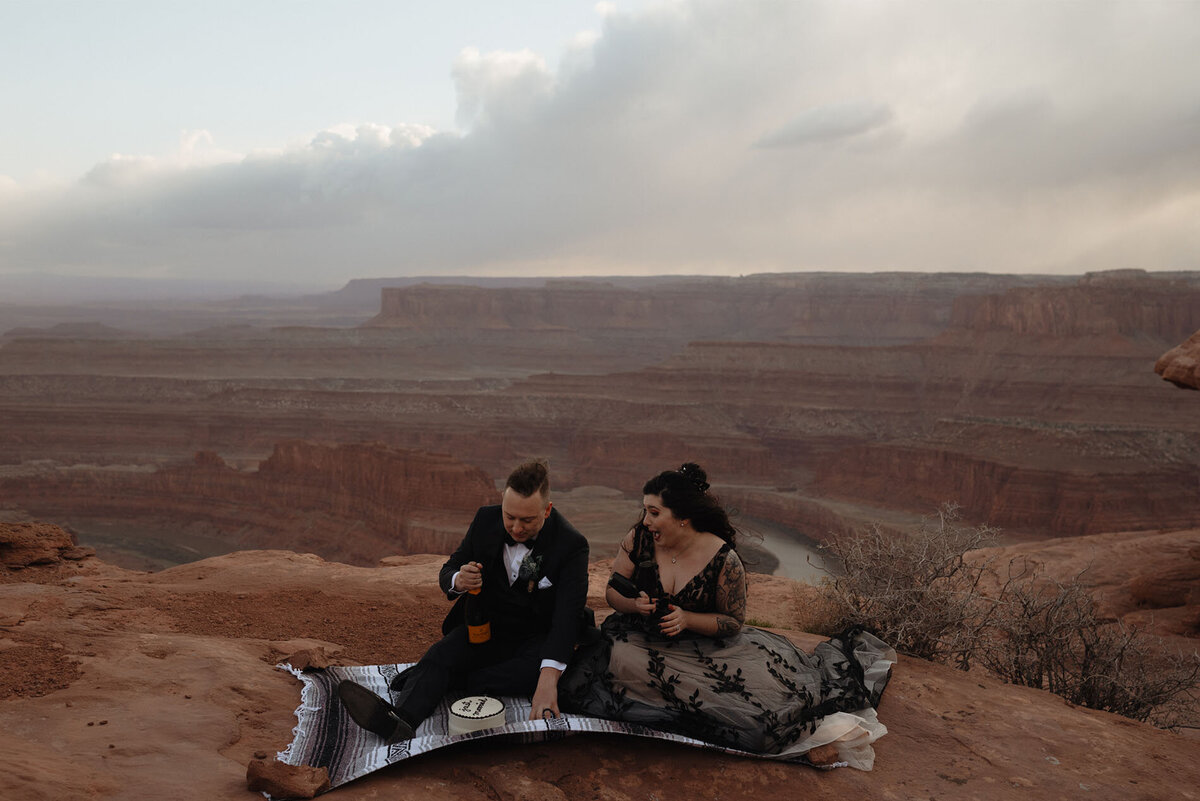 elopement couple sit on a blanket and enjoy champagne and cake at Dead Horse Point State Park in Moab, UT