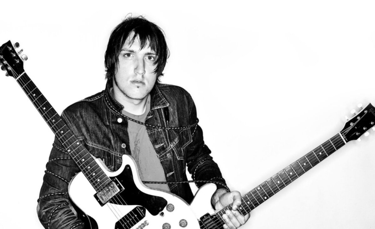 Male musician photo black and white Mikey Manville wearing jean jacket holding two white electric guitars against white backdrop