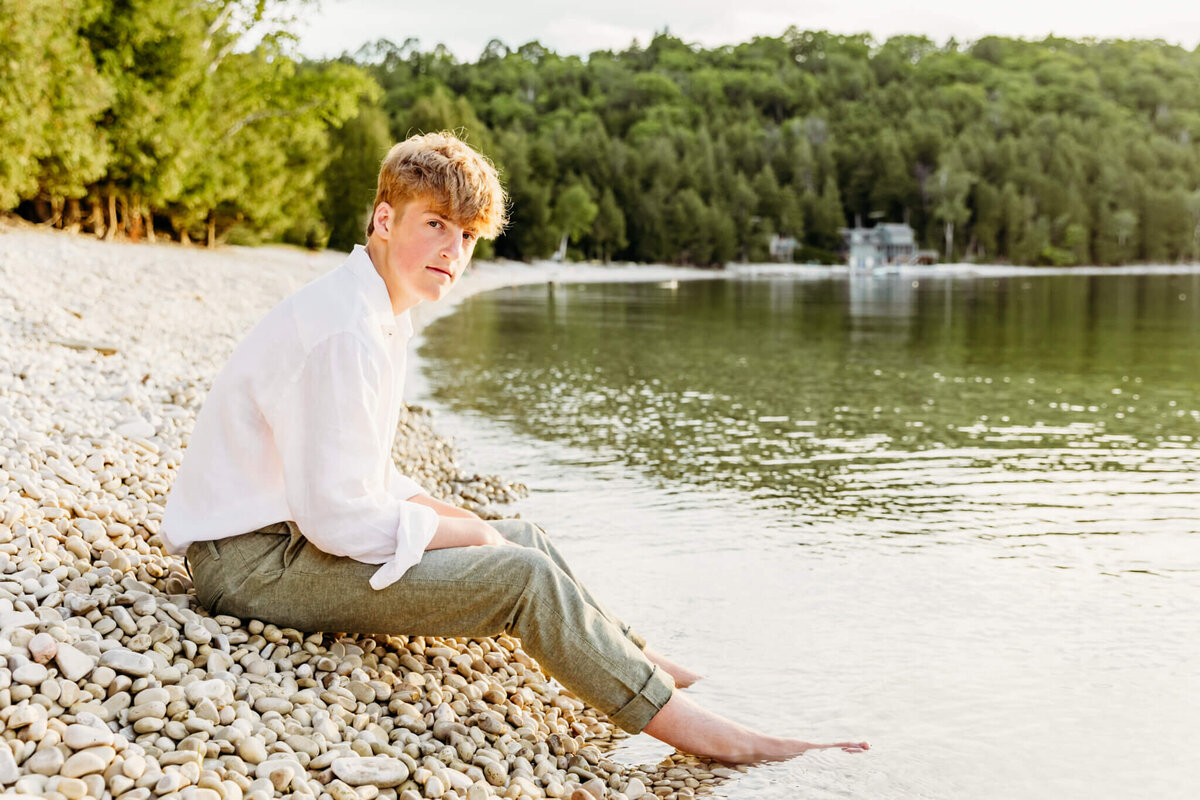 handsome teenage boy in green pants and white button down sitting on a beach with his feet in the water as he looks over he shoulder captured by senior photographer Ashley Kalbus.