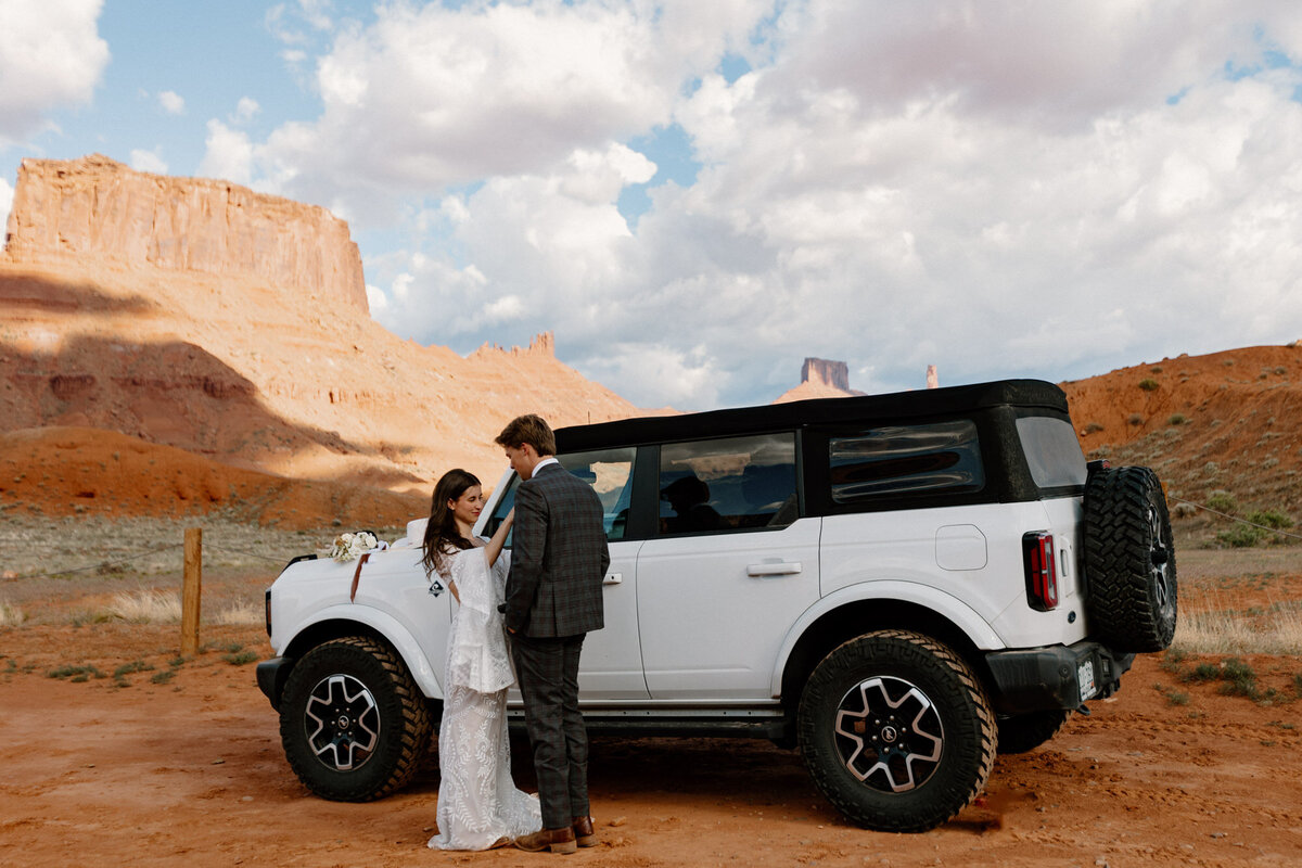 A bride helps her groom get ready for their elopement at Ida Gulch in Mulch. The bride is helping the groom with his Bolo tie, and they are standing in front of their Ford Bronco. 
