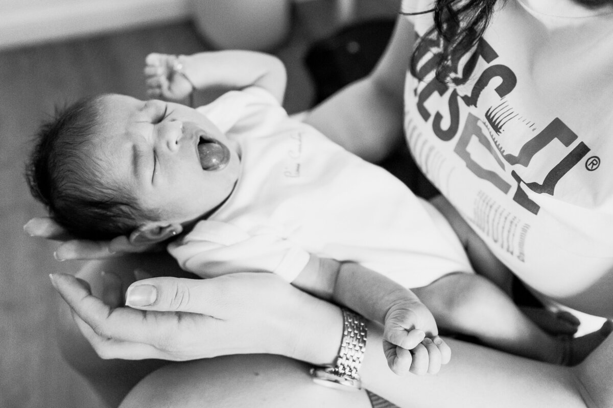 A newborn baby in white onesie yawns widely while supported in the hands of an adult wearing a white t-shirt.