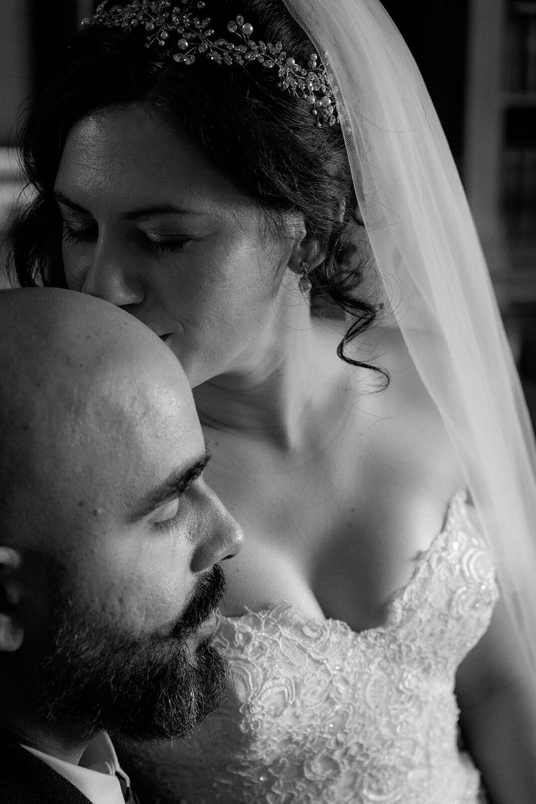 A bride kissed her grooms bald head as he closes his eyes.