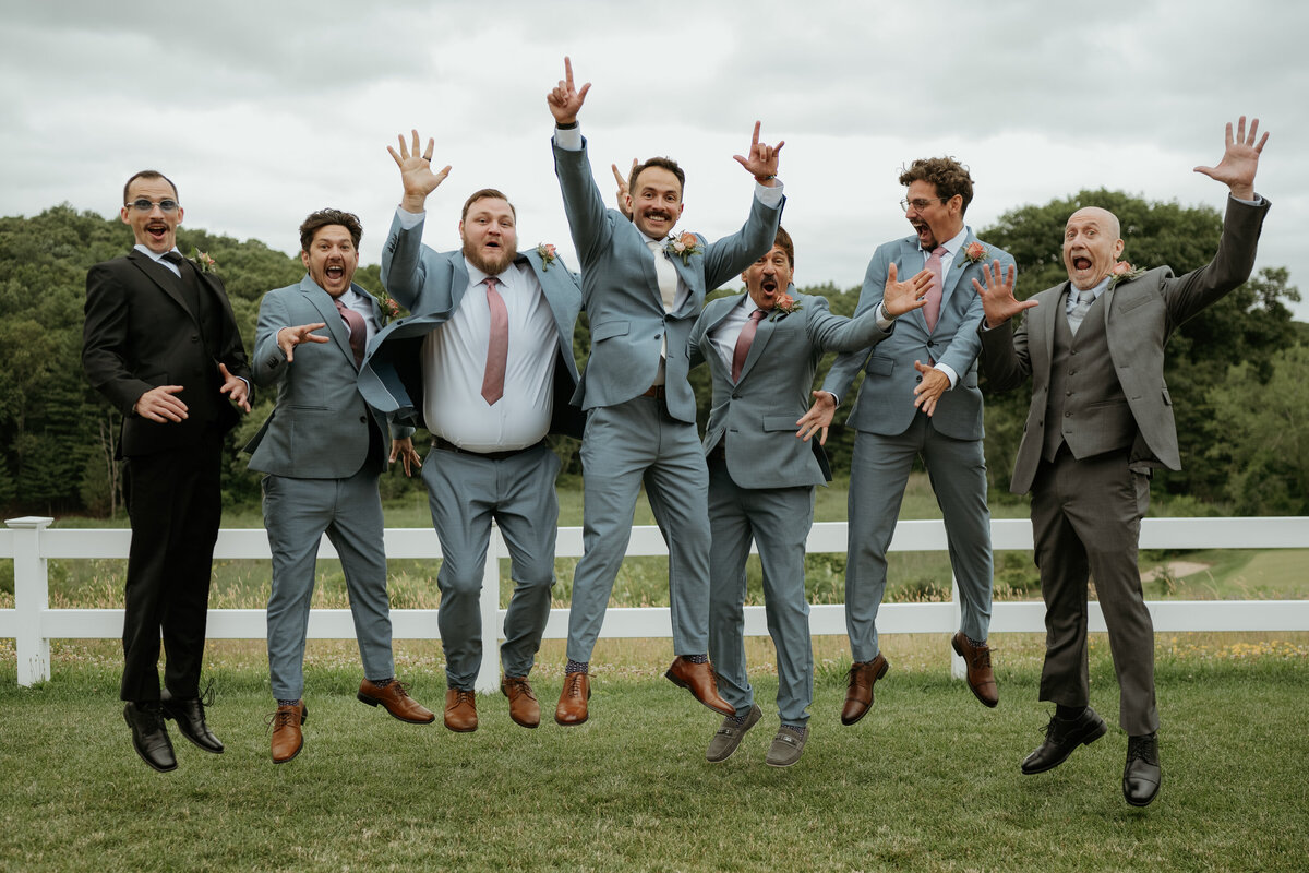 Groomsmen in light blue suits and pink ties jumping in the air with playful expressions in front of a white fence and lush greenery. The groom is at the center, clearly enjoying the moment with his friends.