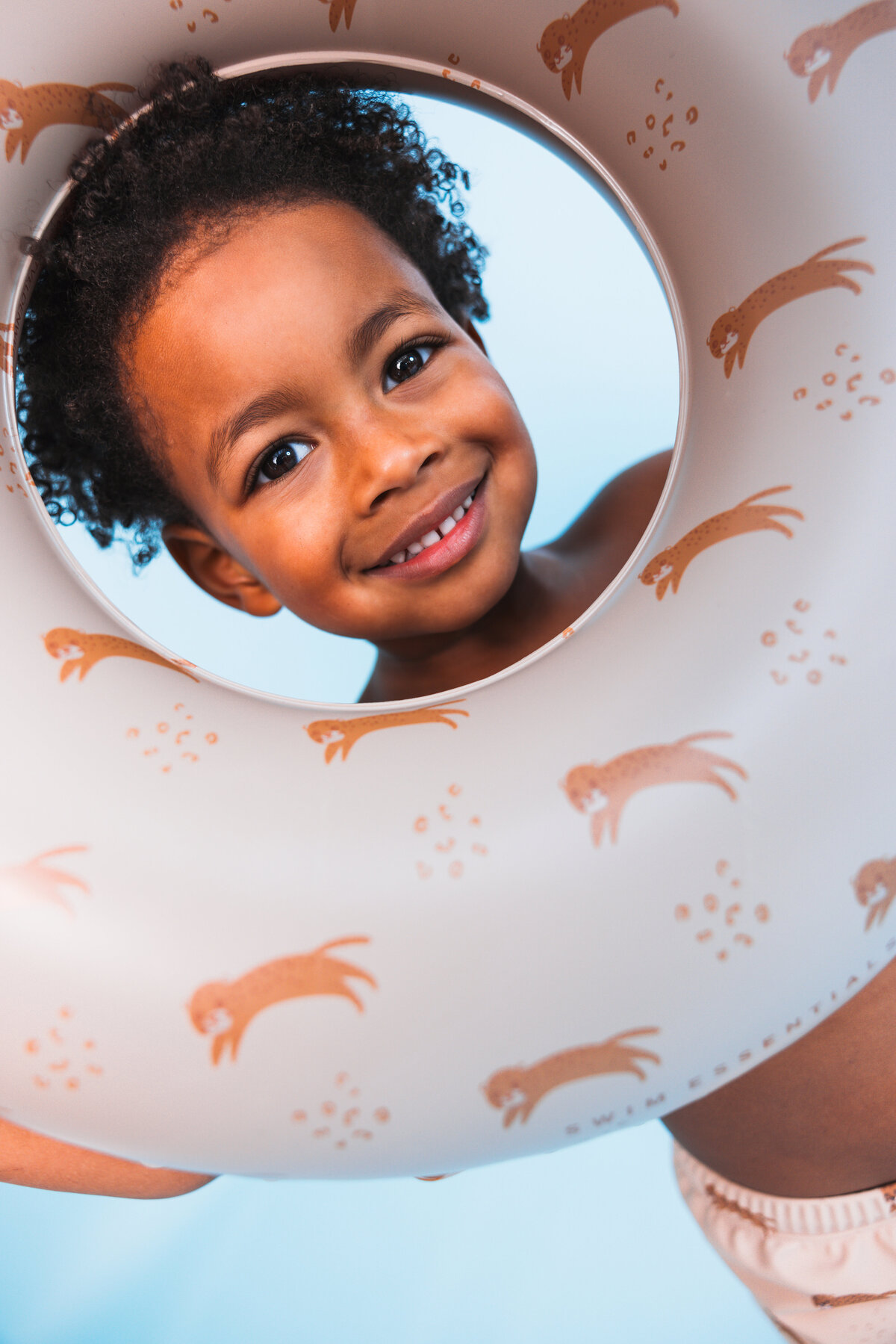 Smiling child looking through a beige Swim Essentials swim ring with playful otter prints, set against a light blue background.