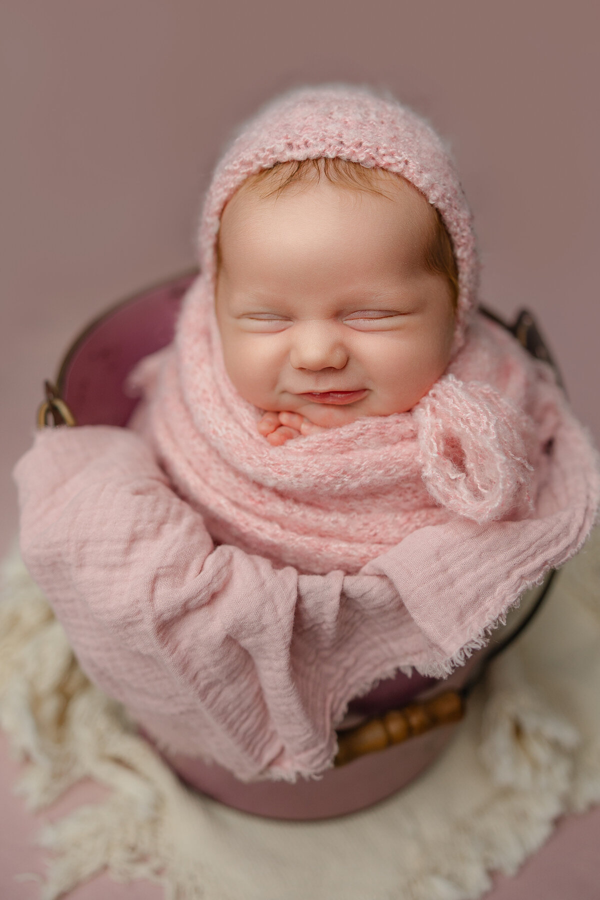 Smiling newborn baby girl wrapped in soft pink knits with a matching bonnet, posed in a bucket during her studio session. Captured by Inland Empire newborn photographer Kaitlyn Dawn Photography.