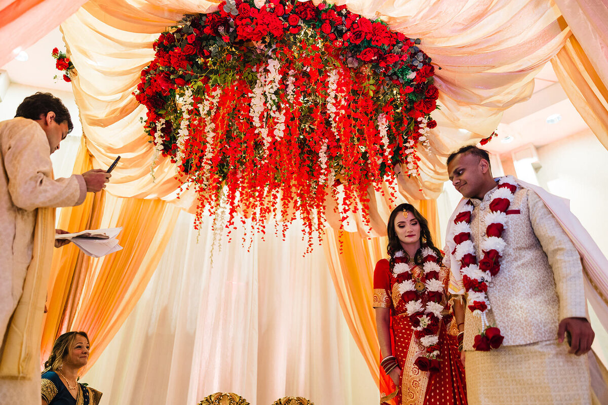 A bride in a red saree and a groom in a cream sherwani standing under a lavish floral arrangement of red and white flowers at their wedding venue.