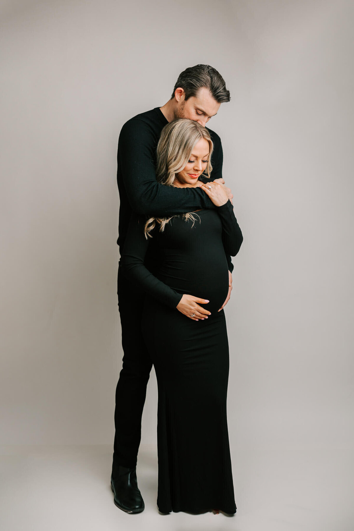 studio photography portrait of pregnant blonde woman wearing black being hugged by husband also wearing head to toe black on white backdrop.