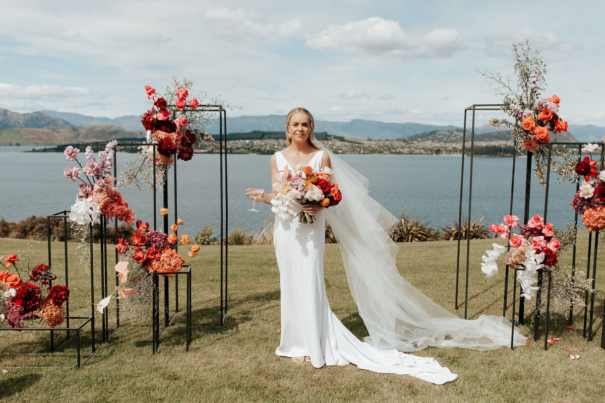 The Vase Floral Co - bride with bouquet stands next to Queenstown lake in between wirframes with floral arrangements