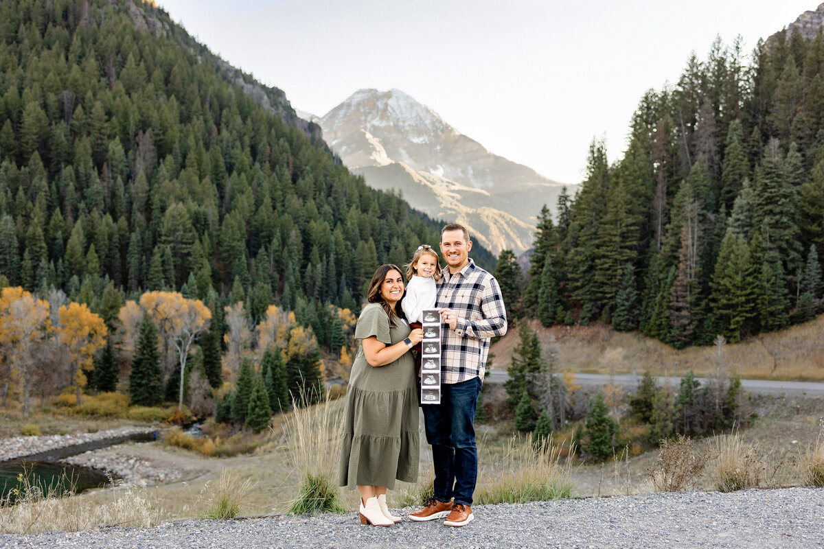 A young family proudly displays ultrasound images as they stand against a breathtaking mountainous landscape, celebrating their upcoming addition with glowing smiles.