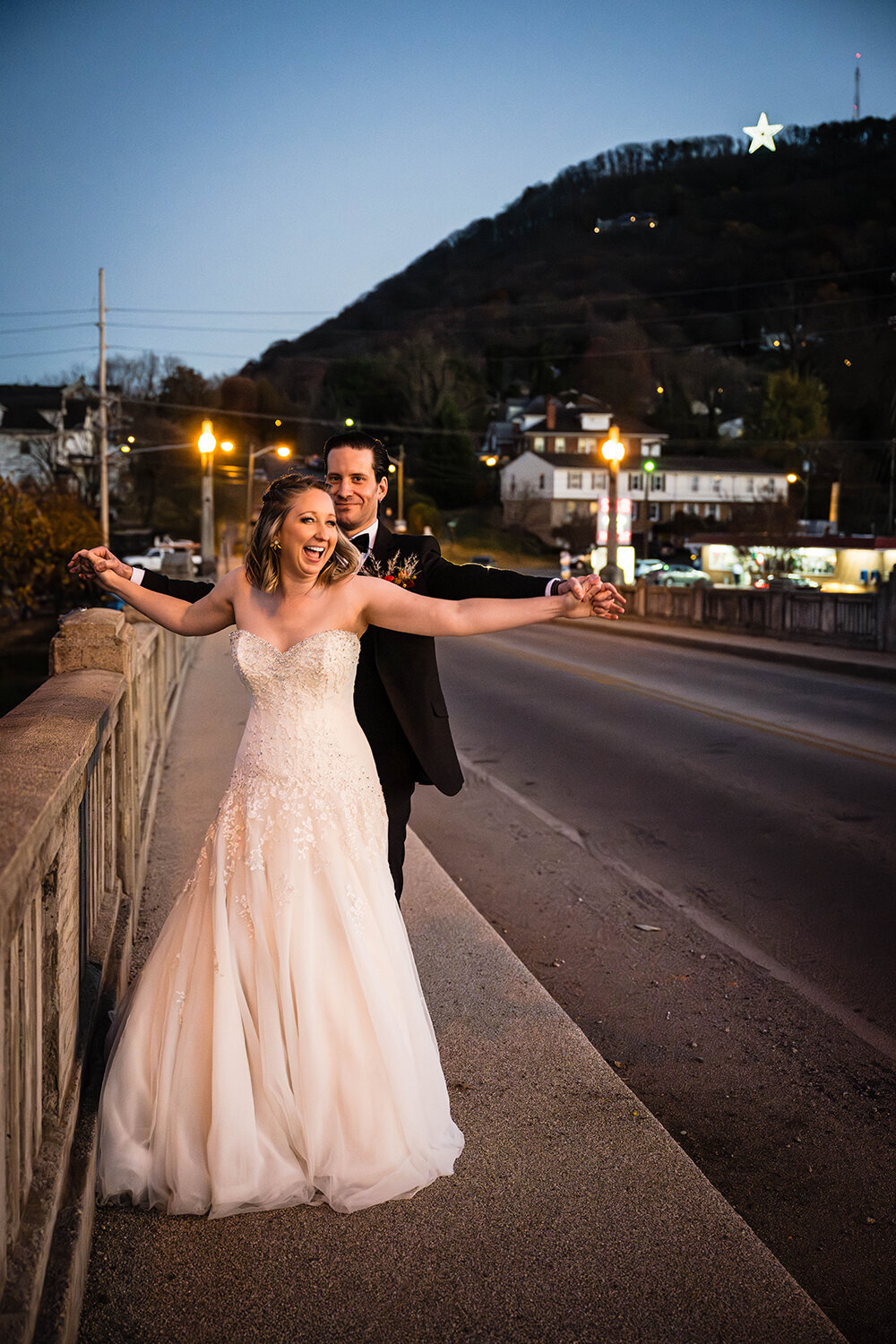 A bride stands in front of a groom while their fingers are intertwined and arms are up in the air moving at the bridge leading up to the Roanoke Star in Virginia. The groom makes a funny face while the bride smiles widely.