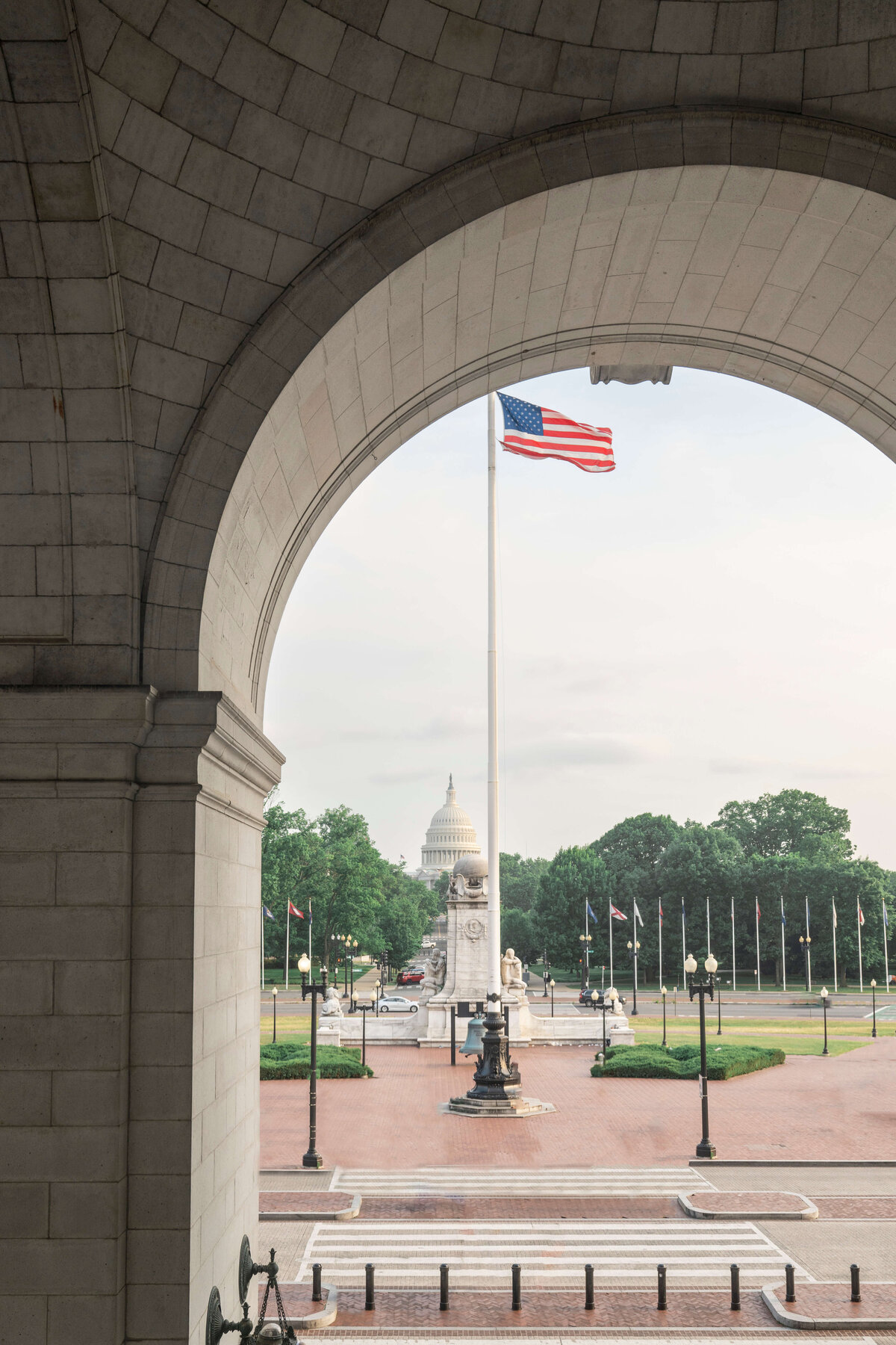 View through an archway of the U.S. Capitol building in the distance, framed by an American flag on a tall flagpole. Trees and additional flags line the open area, with a cloudy sky in the background.