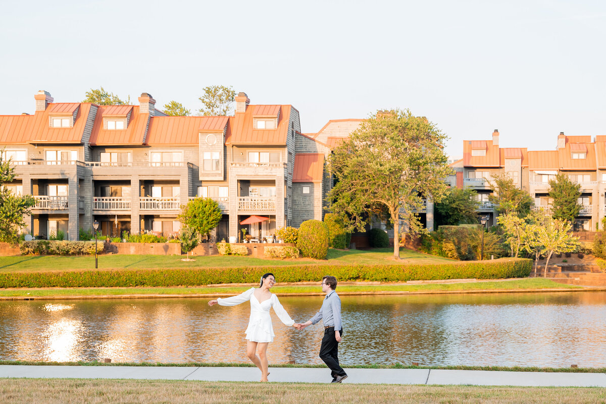 Wide angle image in front of a body of water at the University City Boardwalk where an engaged couple is playfully walking across the frame by Charlotte wedding photographers DeLong Photography