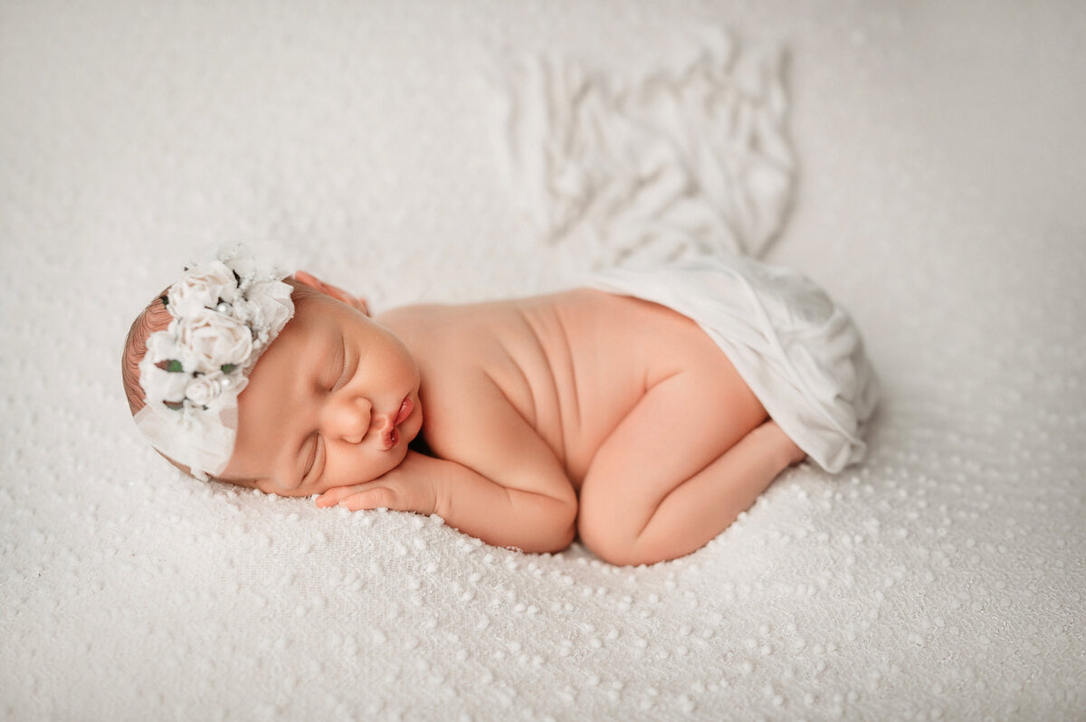 newborn baby girl posing on tummy wearing white floral headband and posed on white textured fabric