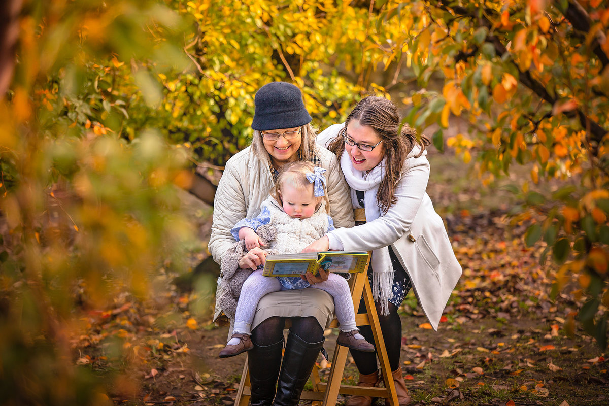 Hampton Roads family photography with generational extended family with grandmother sitting on a stool and reading a book to her granddaughter as the mother of the child leans over and points at the pictures in the book