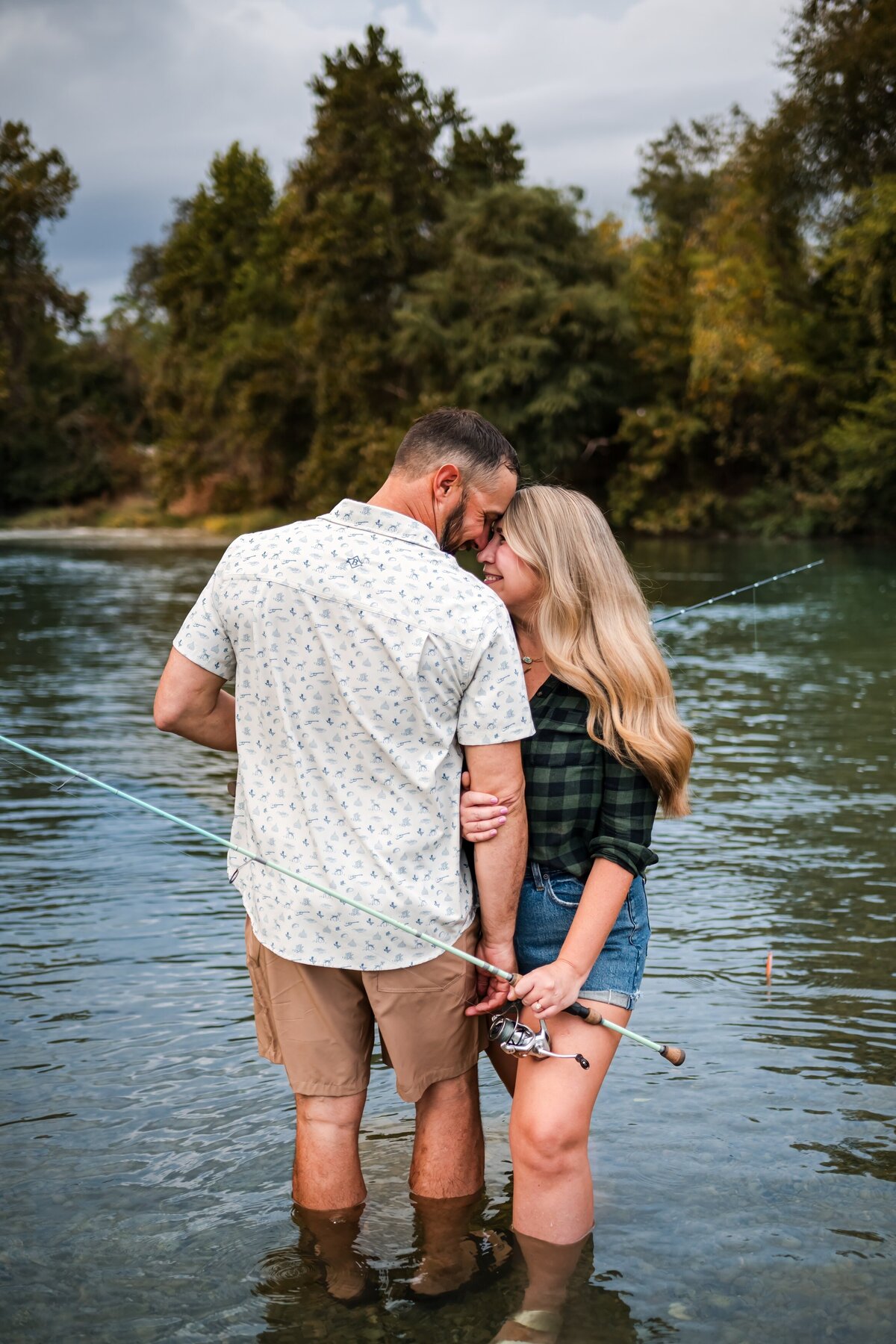 Engaged couple standing side by side with fishing poles, foreheads touching, in the Guadalupe River during lifestyle engagement session in New Braunfels, Texas. The groom faces away while the bride faces the camera."
