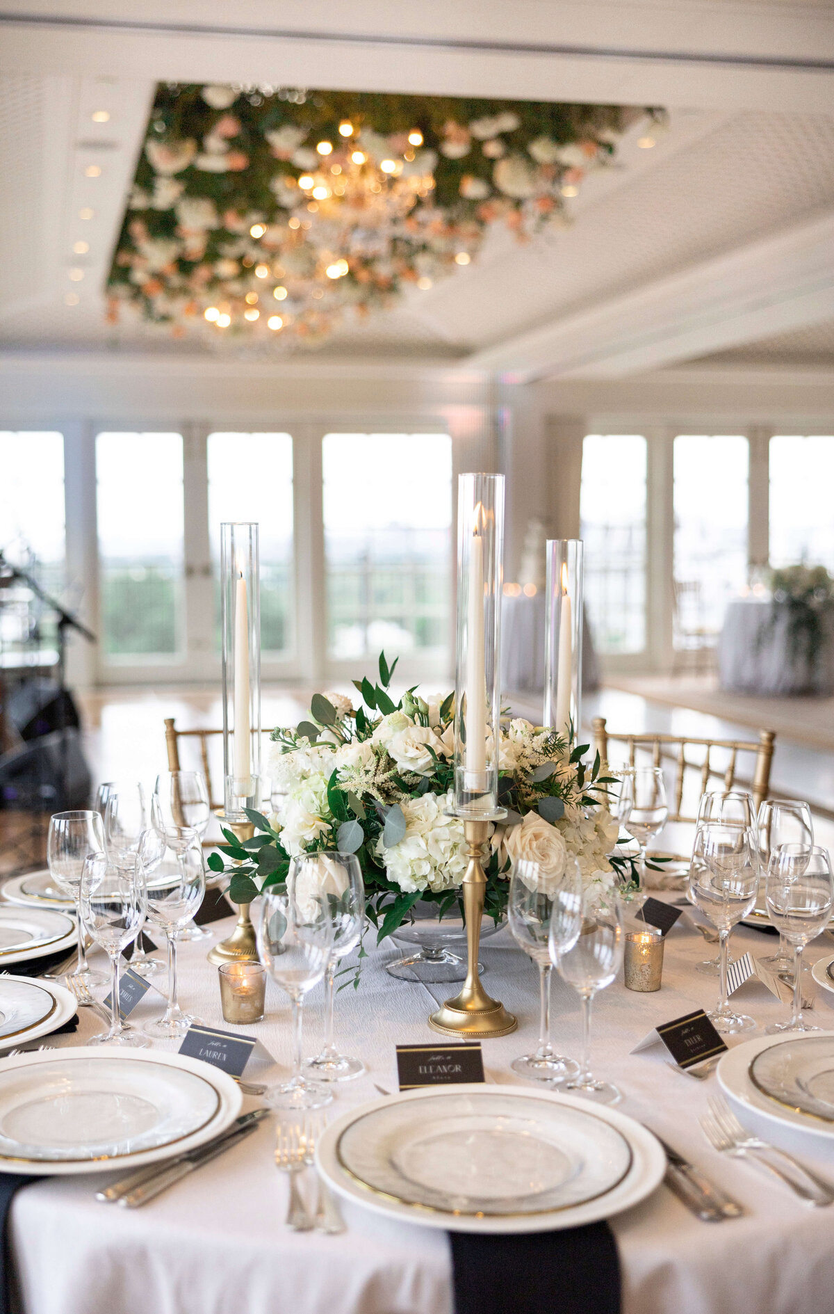 Elegant table setting in a bright, spacious room. A centerpiece of white flowers and greenery is surrounded by tall, lit candles. Gold-rimmed plates, crystal glassware, and neatly placed table settings complete the sophisticated ambiance.