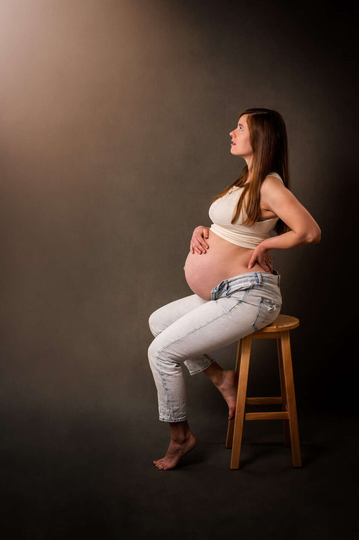 A mam to be in jeans and a white tank top sits on a stool and stares off to the side during her portrait session with an Asheville maternity photographer