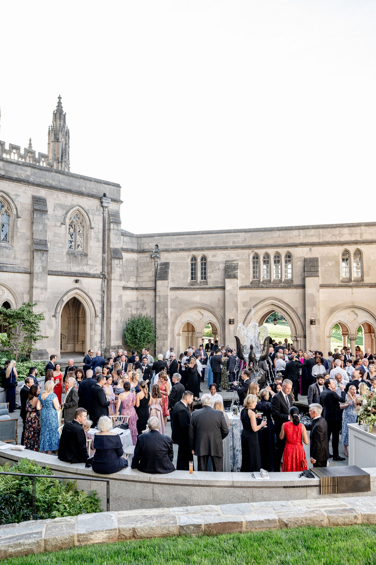 This photo was taken at Washington National Cathedral in Washington D.C. by photographer Twah Dougherty. In the foreground, a large group of people, elegantly dressed in formal attire, including tuxedos and evening gowns, is gathered in the courtyard of a historic stone building with arched doorways and large windows. They are engaged in socializing and celebrating outdoors. Additionally, Current Films served as the videographers capturing the moments of the event.
