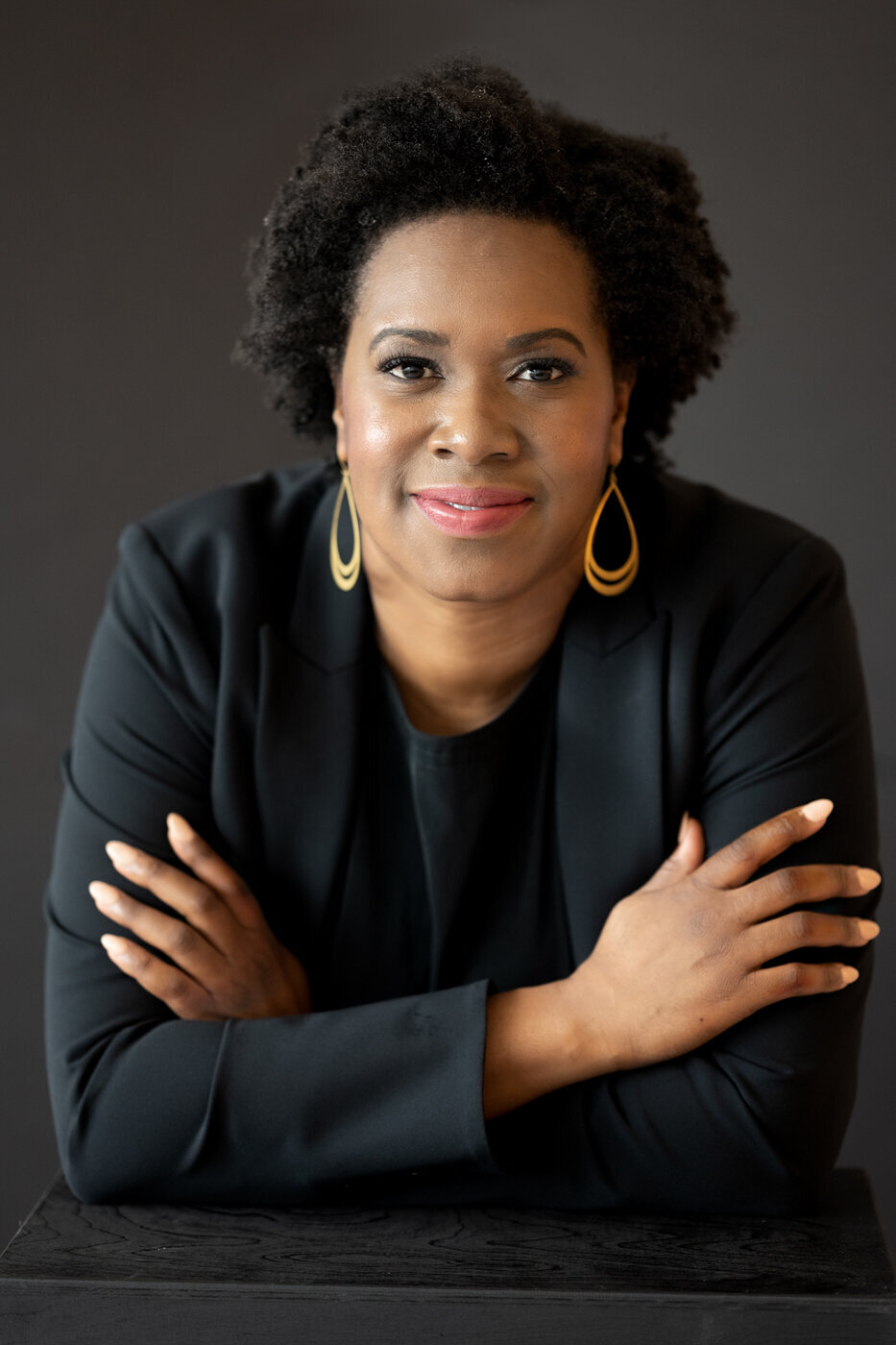 A headshot of a woman wearing black clothes and gold earrings