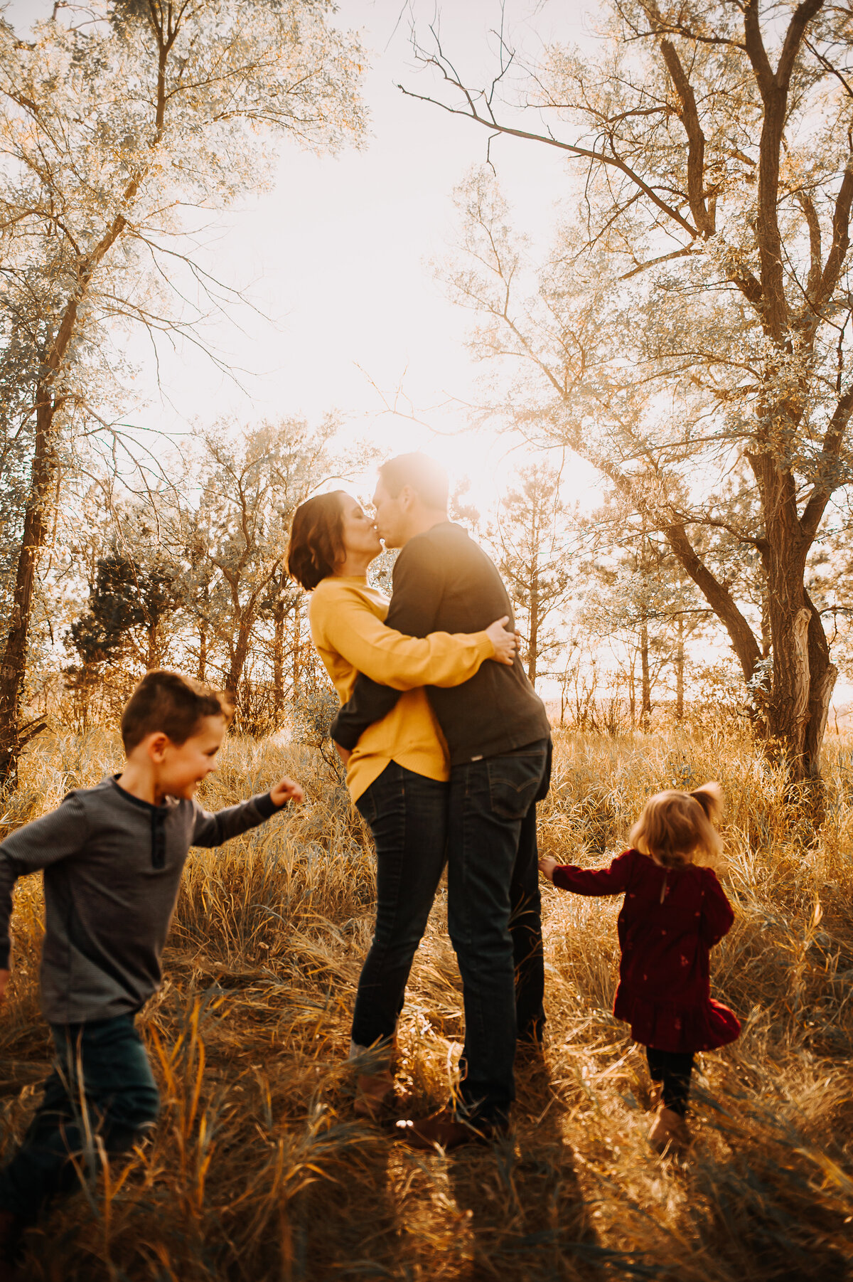 Parents embrace with a little snuggle and a kiss as their young children run circles around them during their fall family mini session at sweet briar lake, nd. 