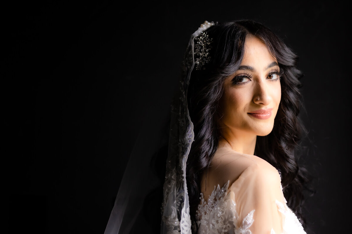 Bride looks at camera and slightly smiles.  The background is black and you can see her face, dress and veil beautifully.