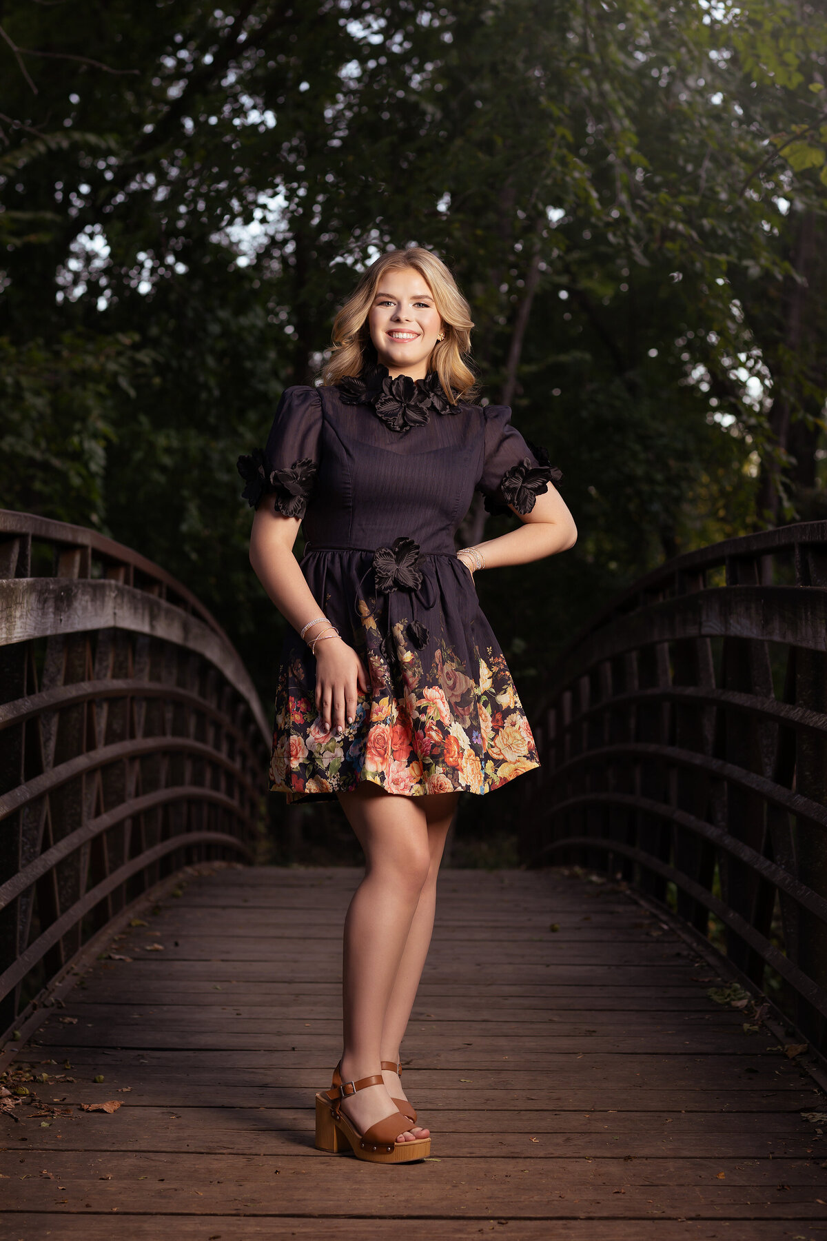 Dallas senior photography of a young woman in a floral dress, standing on a wooden bridge in nature.