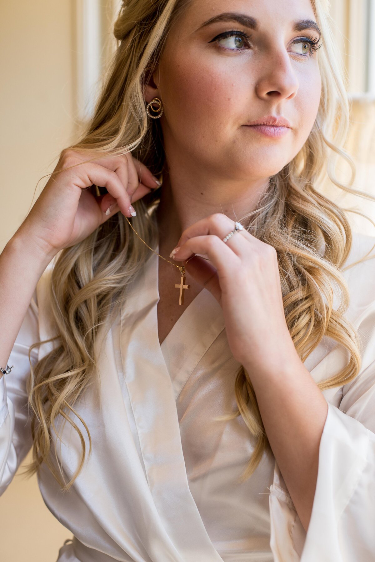 a blond bride adjusts a necklace as she gets prepared for her wedding day. Her hair is softly curled and she wears a white robe