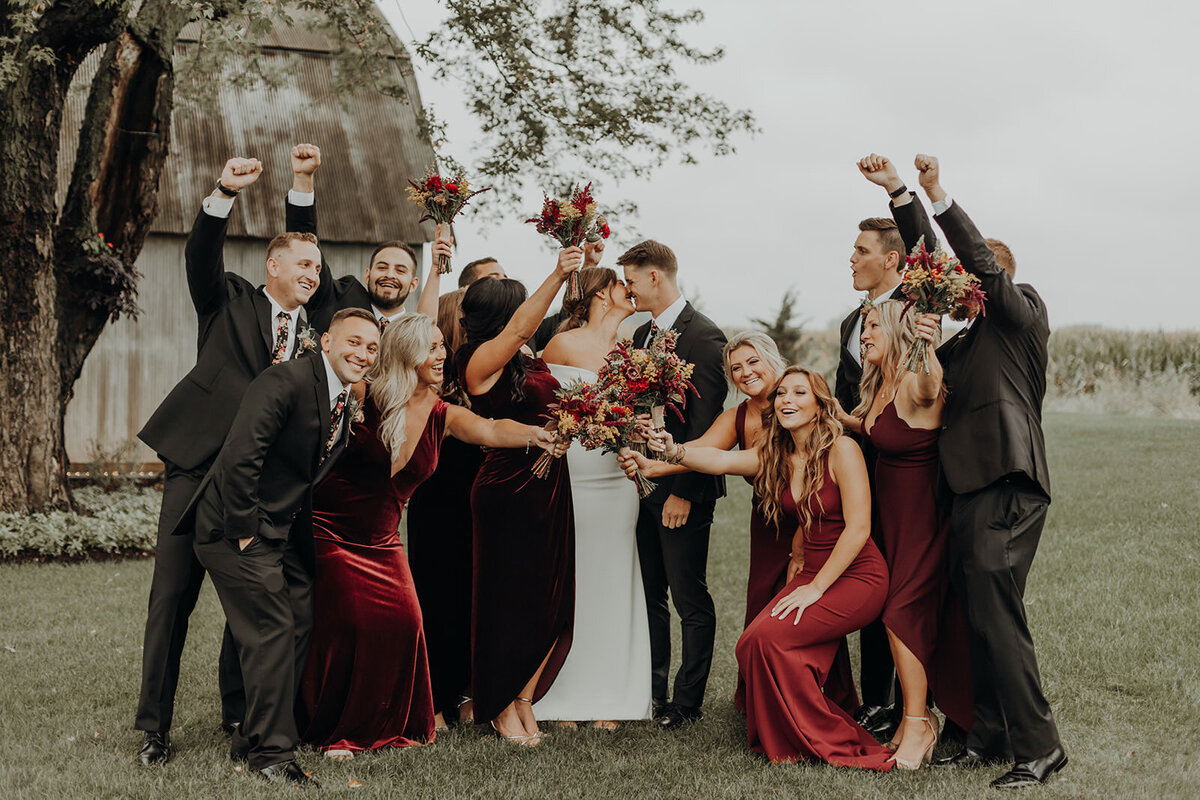 Bride and groom kiss while surrounded by their cheering wedding parties