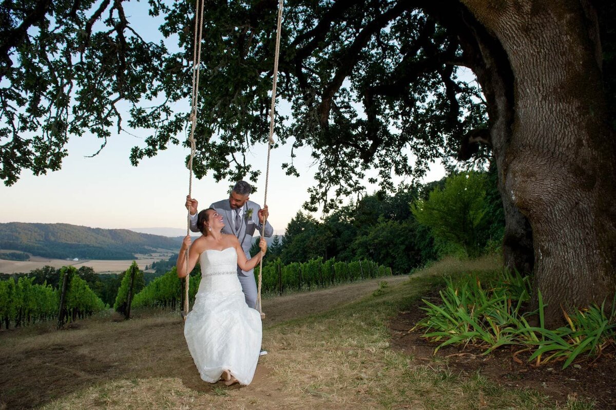 a man pushes his bride in a rope swing hanging from an old oak tree overlooking a vineyard