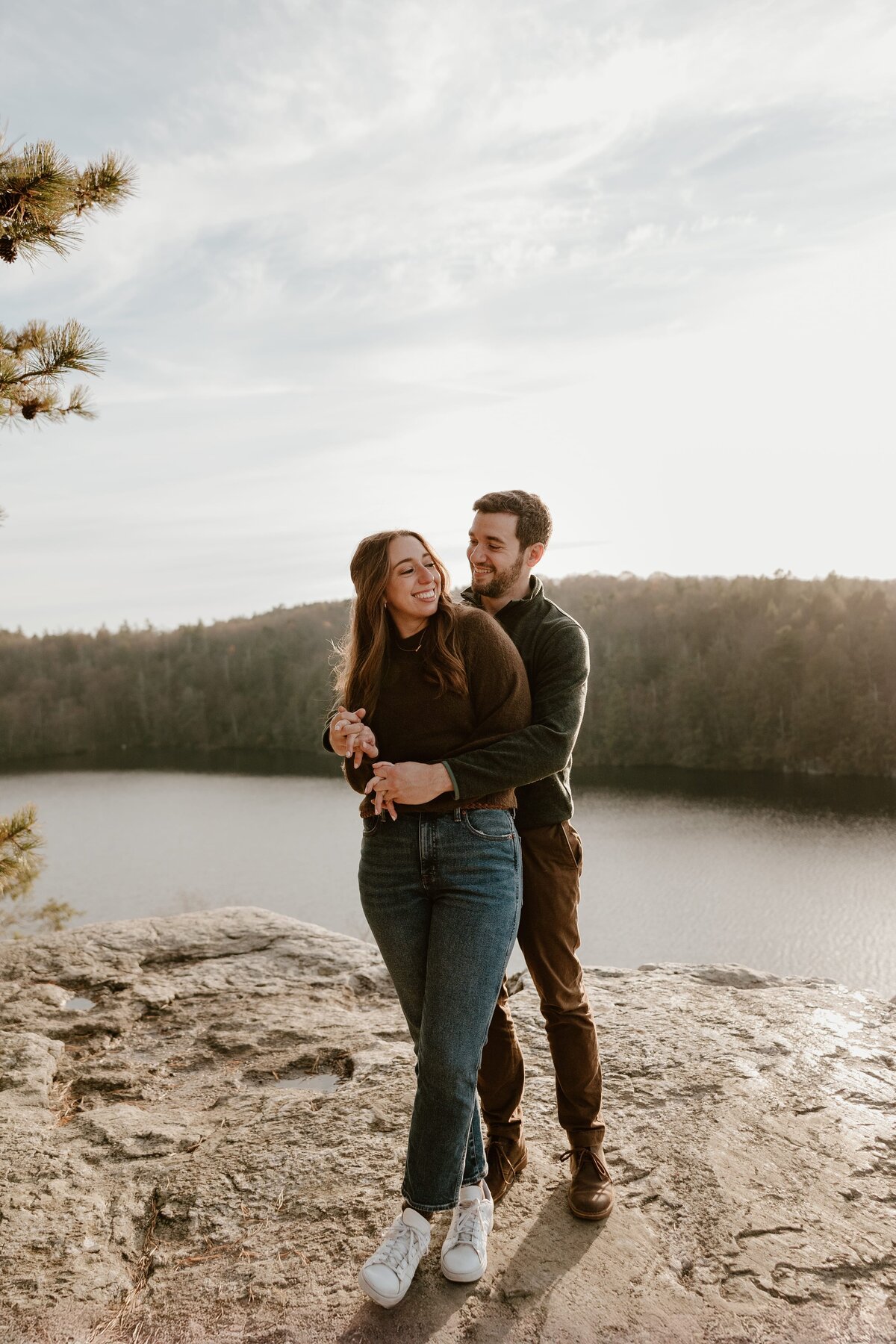 Engaged couple sharing a joyful embrace on a rocky overlook at Lake Minnewaska, surrounded by a peaceful view of the lake and forest in the background under a soft, cloudy sky.