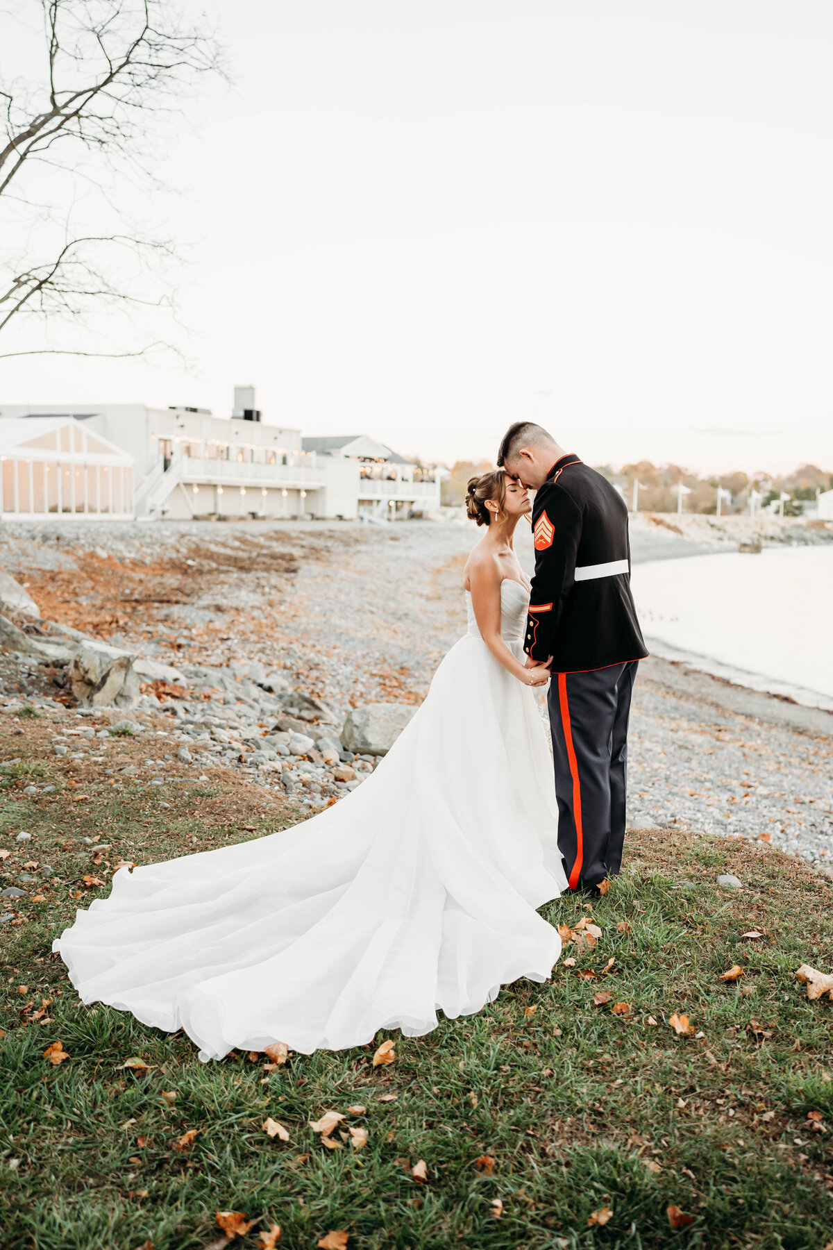 Romantic waterfront wedding at Oceanview of Nahant, MA. A stunning bride in a flowing strapless gown shares an intimate moment with her groom in a U.S. Marine dress uniform against a picturesque rocky shoreline at sunset. Captured by Captured by Moll, a Boston wedding photographer specializing in timeless coastal wedding photography. Perfect inspiration for military weddings, beachside ceremonies, and New England wedding venues.