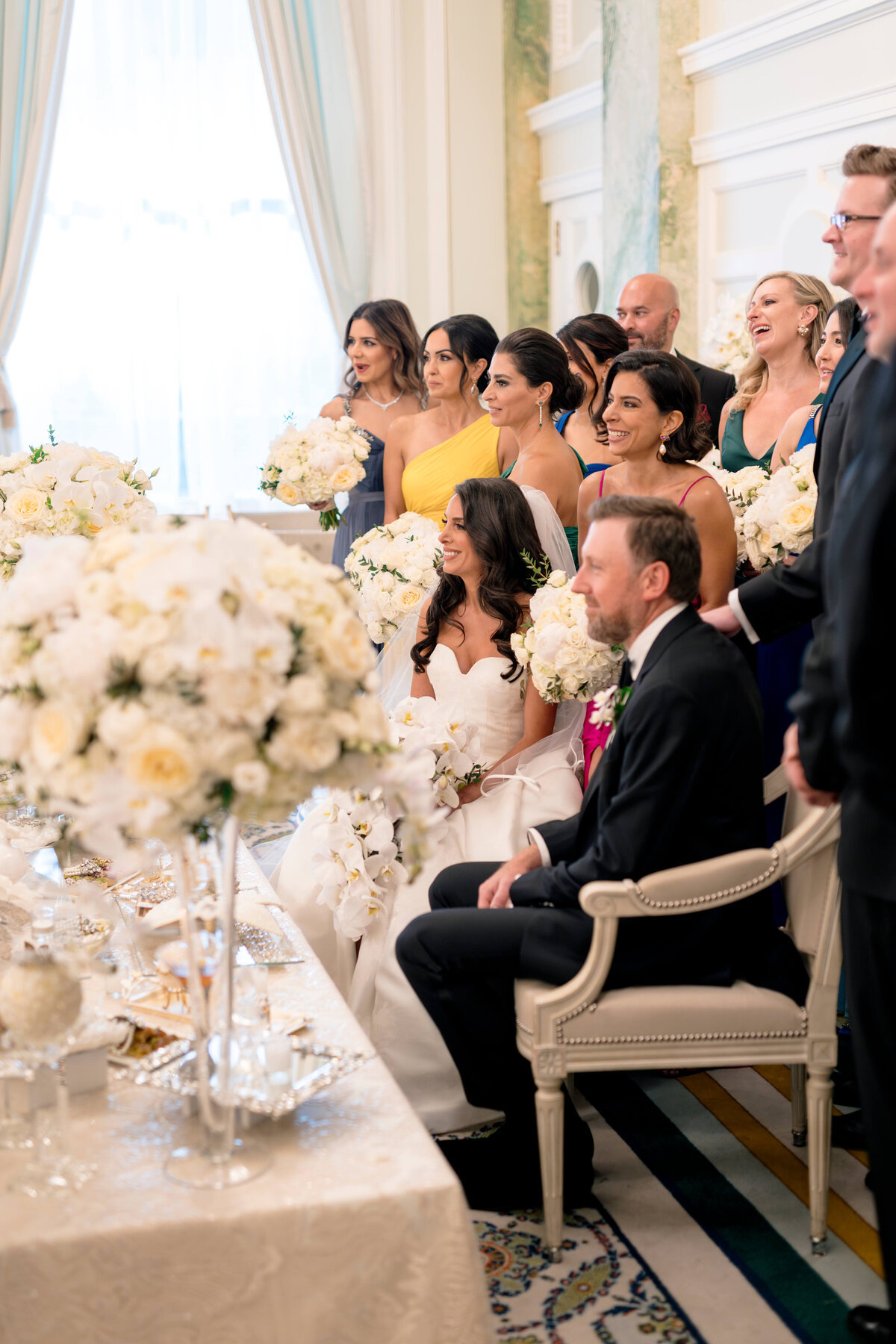 A bride and groom sit side by side, surrounded by a group of smiling bridesmaids and groomsmen. The bridesmaids hold white bouquets and wear dresses in shades of yellow and blue. The room is elegantly decorated with floral arrangements.
