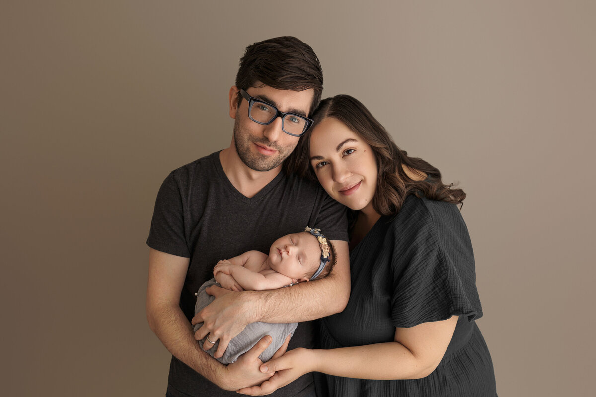 Parents holding their sleeping newborn baby wrapped in a grey blanket, posing against a neutral beige background, both parents smiling warmly.