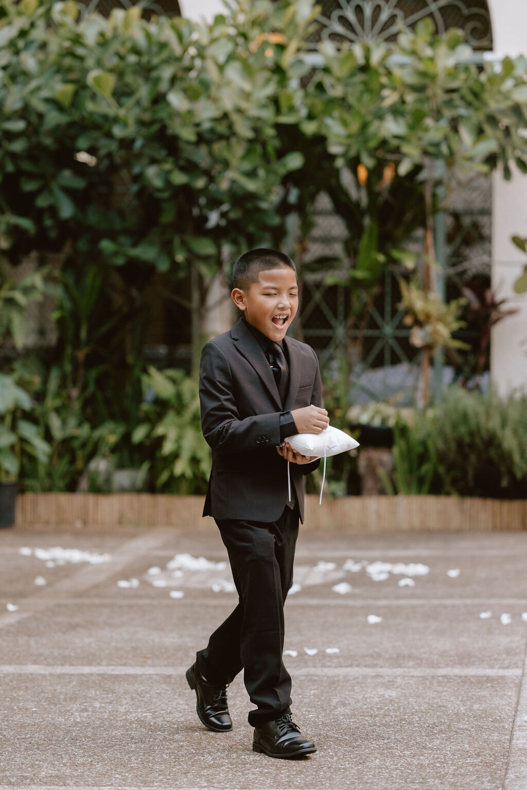 Ring bearer in an all black suit with greenery behind him looking very excited
